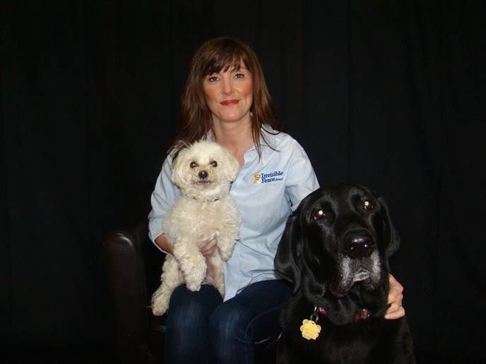 A person sits in front of a black backdrop, holding a small white dog while a large black dog sits beside them.