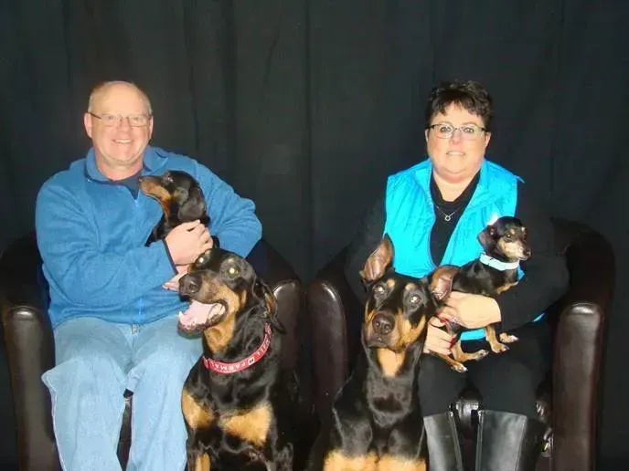 A man and a woman sit in chairs, each holding a small dog, with two large dogs sitting on the floor in front of them.
