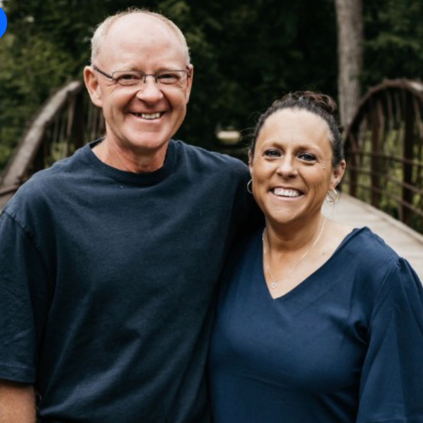 A man and woman in blue shirts smiling for a photo together on a wooden bridge outdoors.