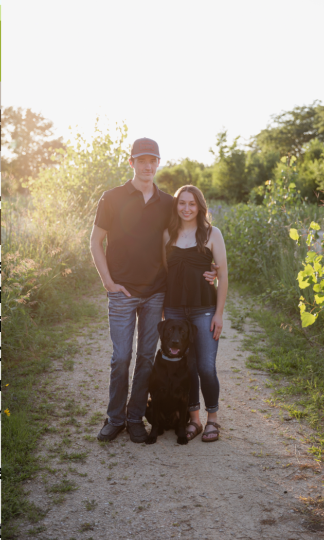 A student in a maroon cap and gown sits on a ledge in front of a brick campus building, holding their graduation cap.