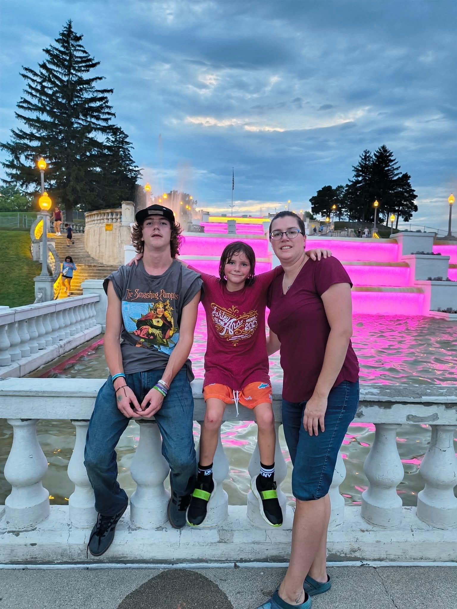 Three people pose on a stone railing in front of a multi-tiered fountain illuminated with bright pink lights at dusk.
