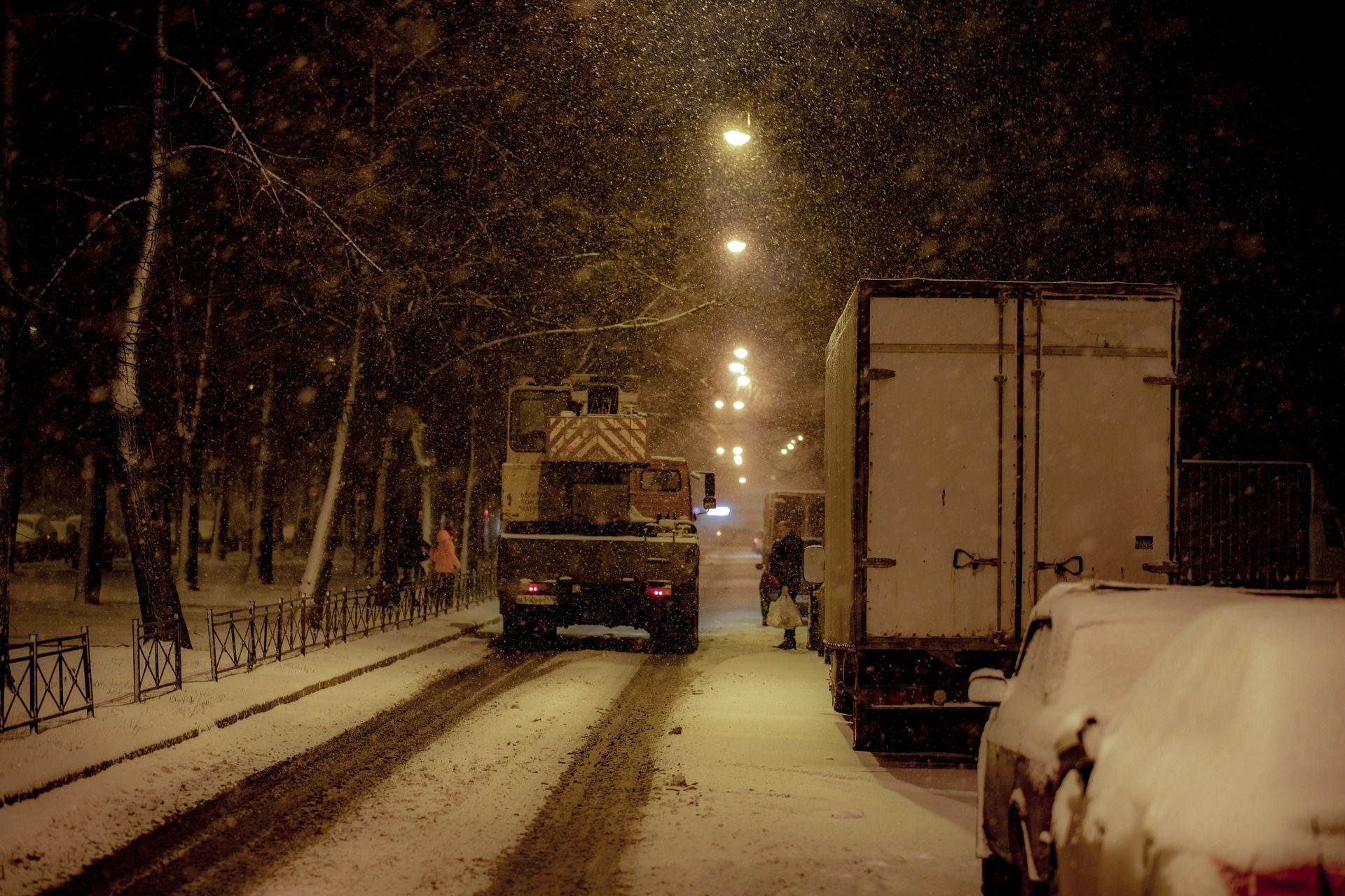 Snowy night scene: truck plowing a snow-covered street lined with trees and parked vehicles.