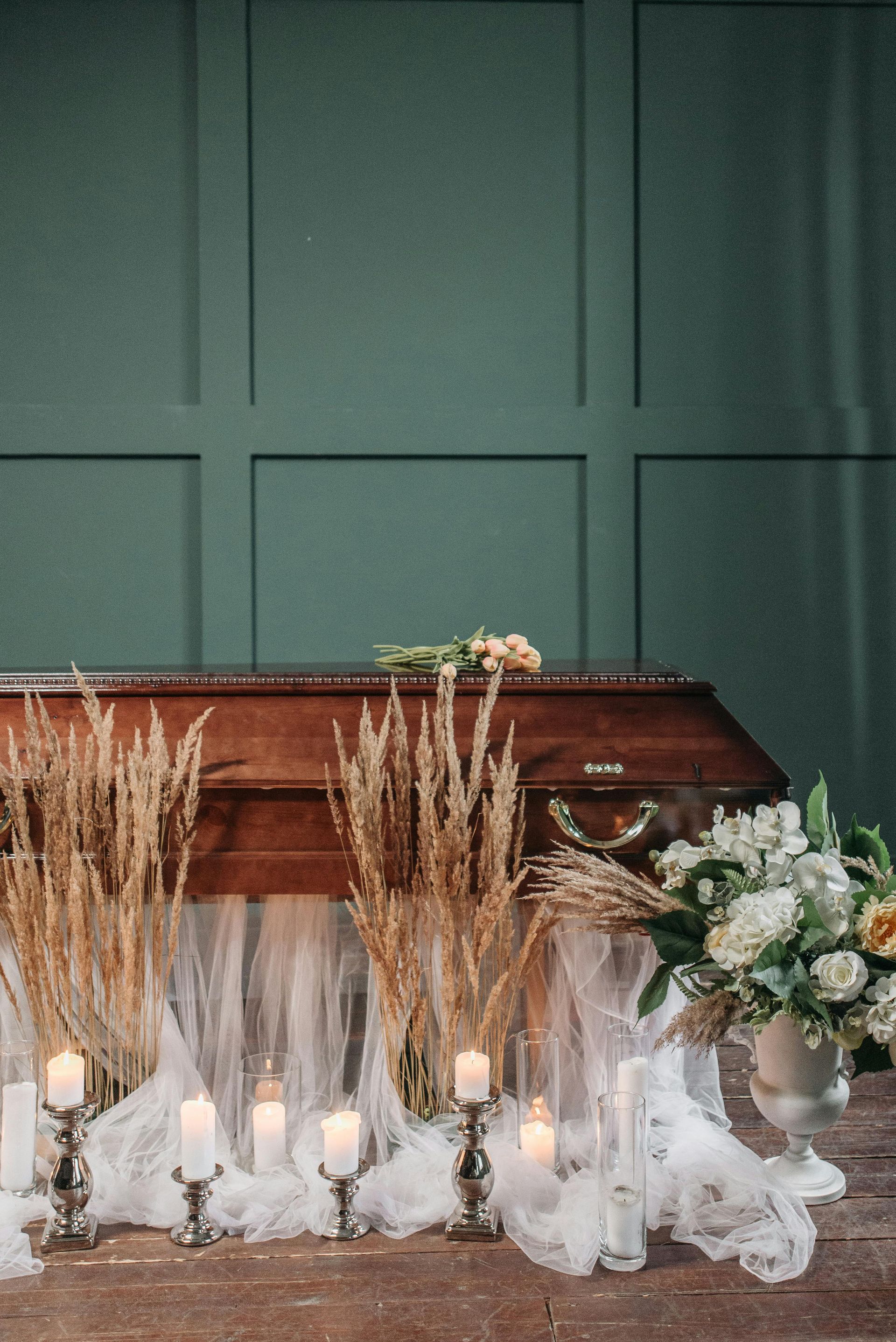 Brown casket decorated with flowers and candles in front of a green paneled wall.