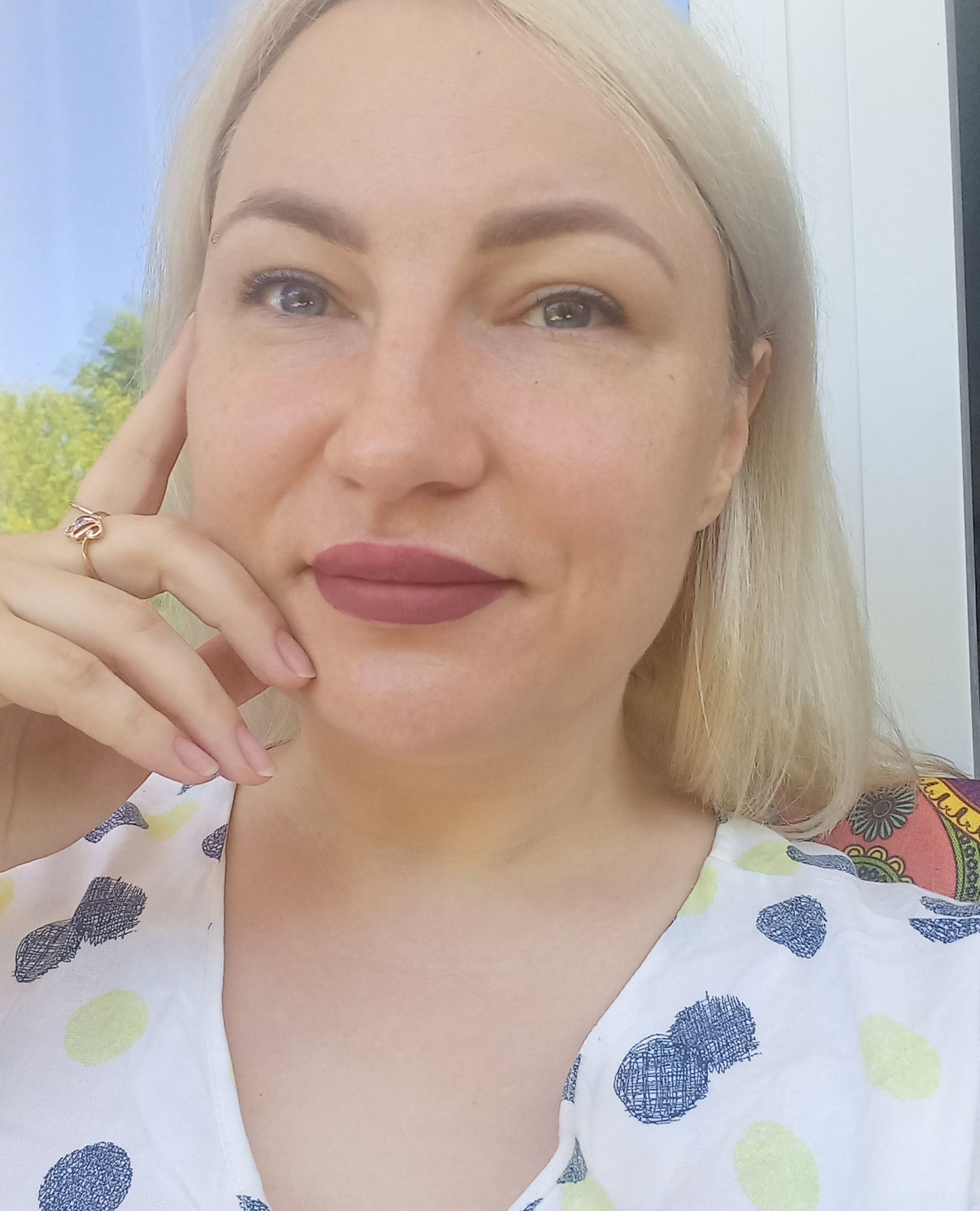 A blonde person with a neutral expression poses with their hand near their face, wearing a white pineapple-print top.