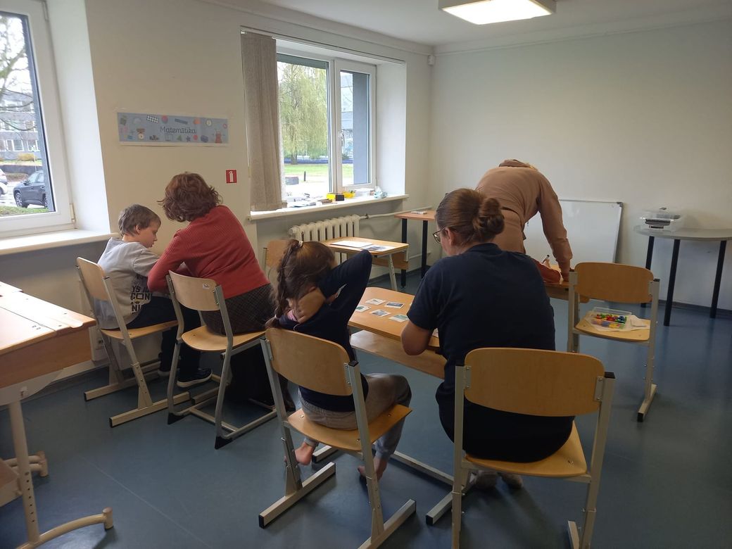 Two children and two adults work at tables in a bright, modern classroom.