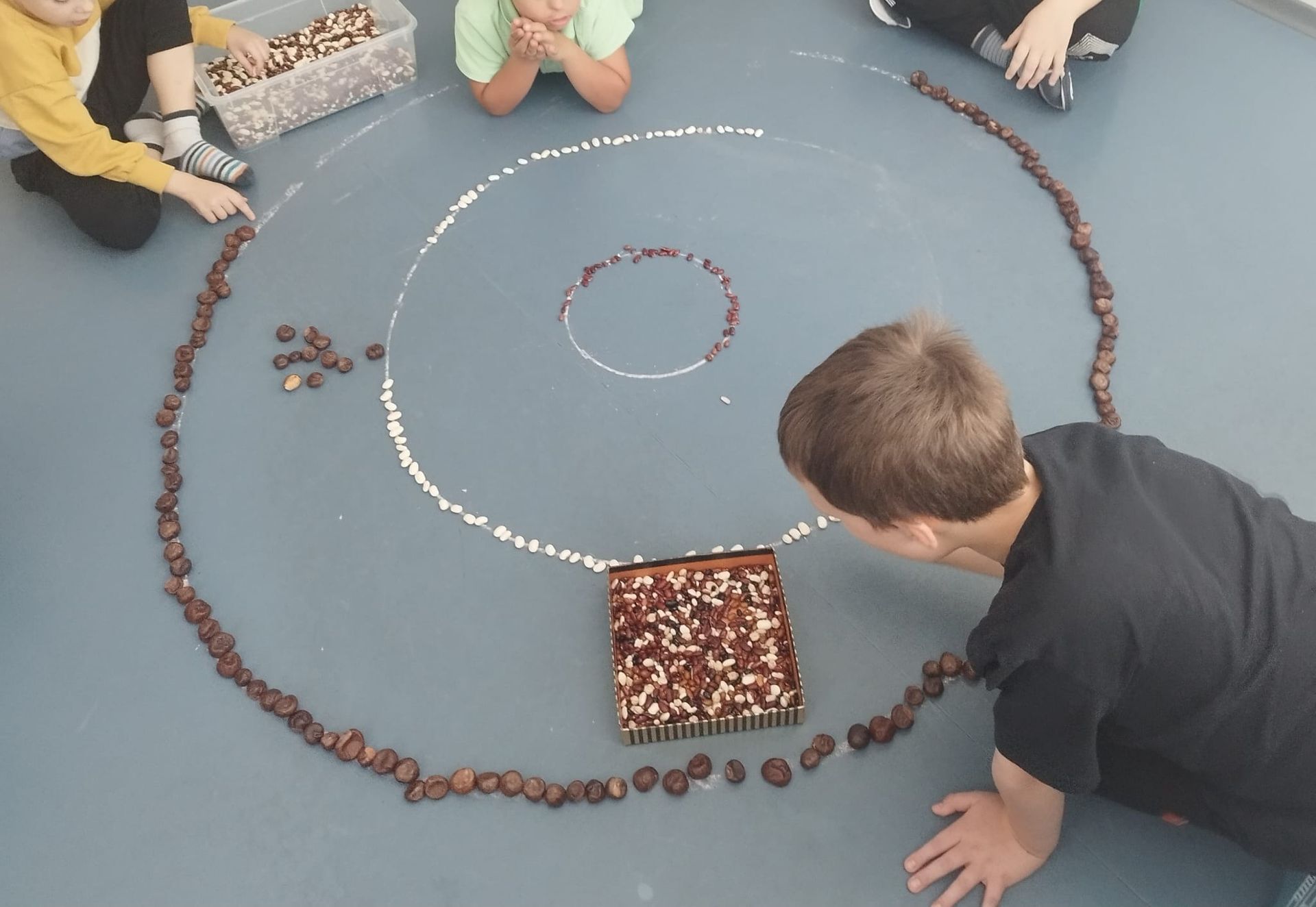 Children arranging chestnuts and white beads in concentric circles on a blue floor.