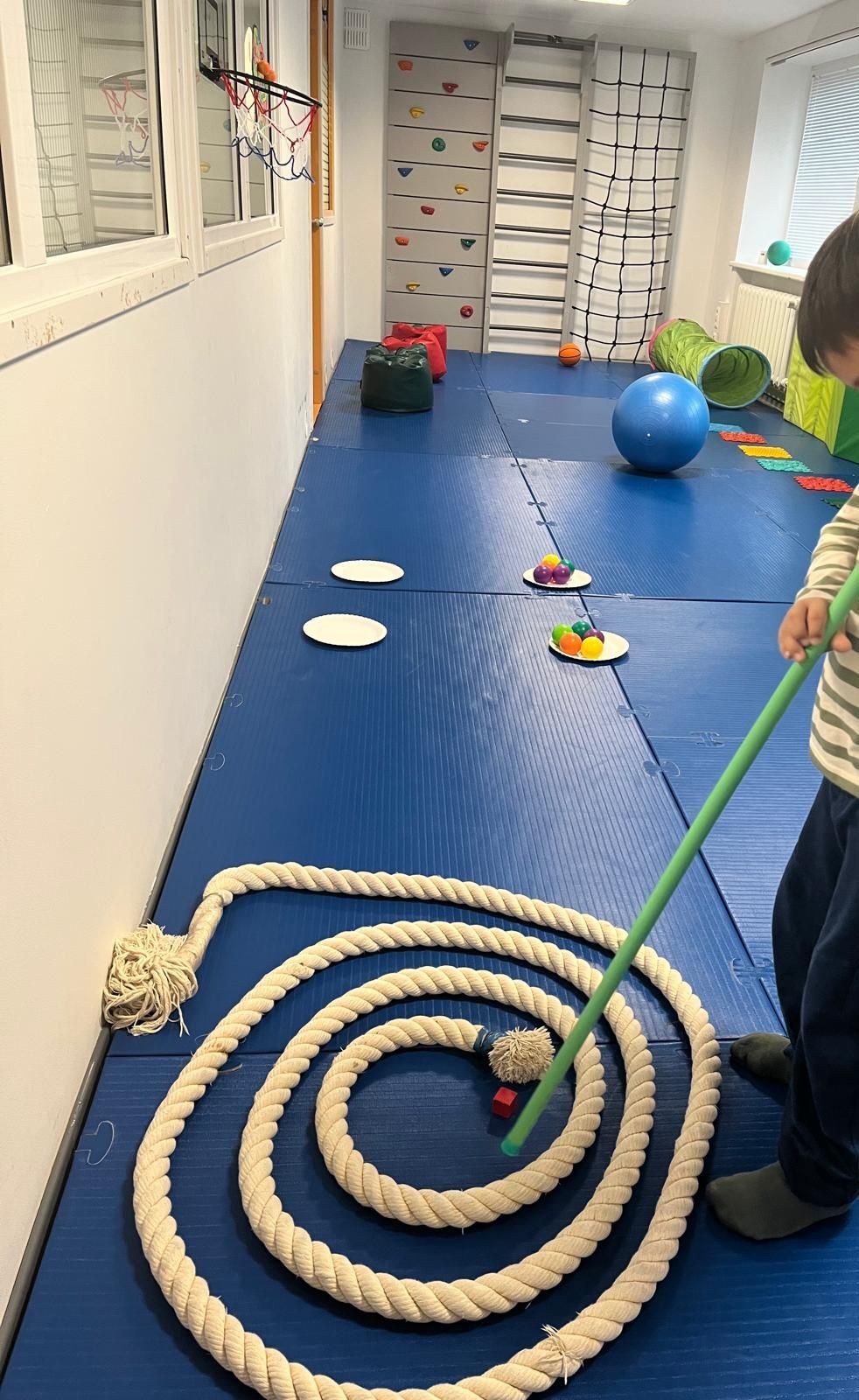 Child using a stick to guide a ball through a coiled rope on a blue mat, in a playroom with climbing walls.