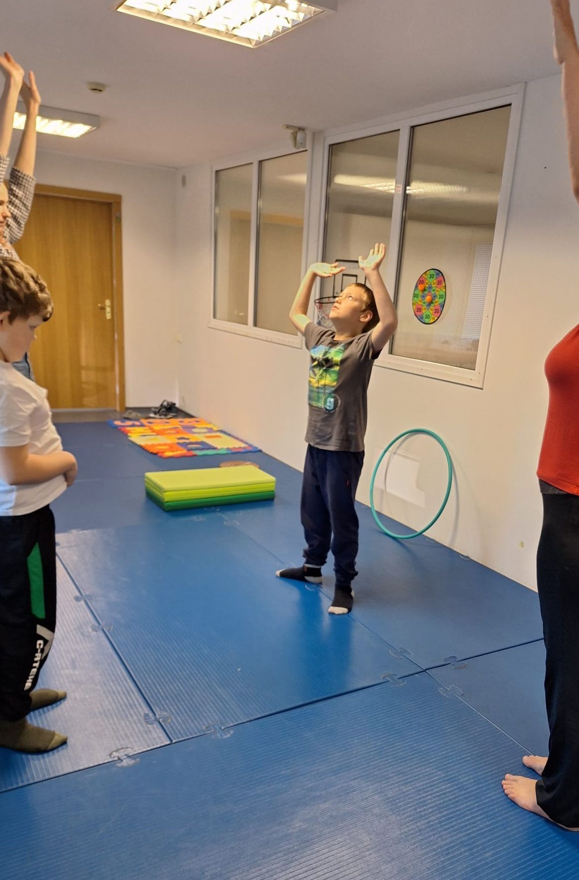 A young person in a gray shirt raises their arms in a room with others. Blue floor, white walls.
