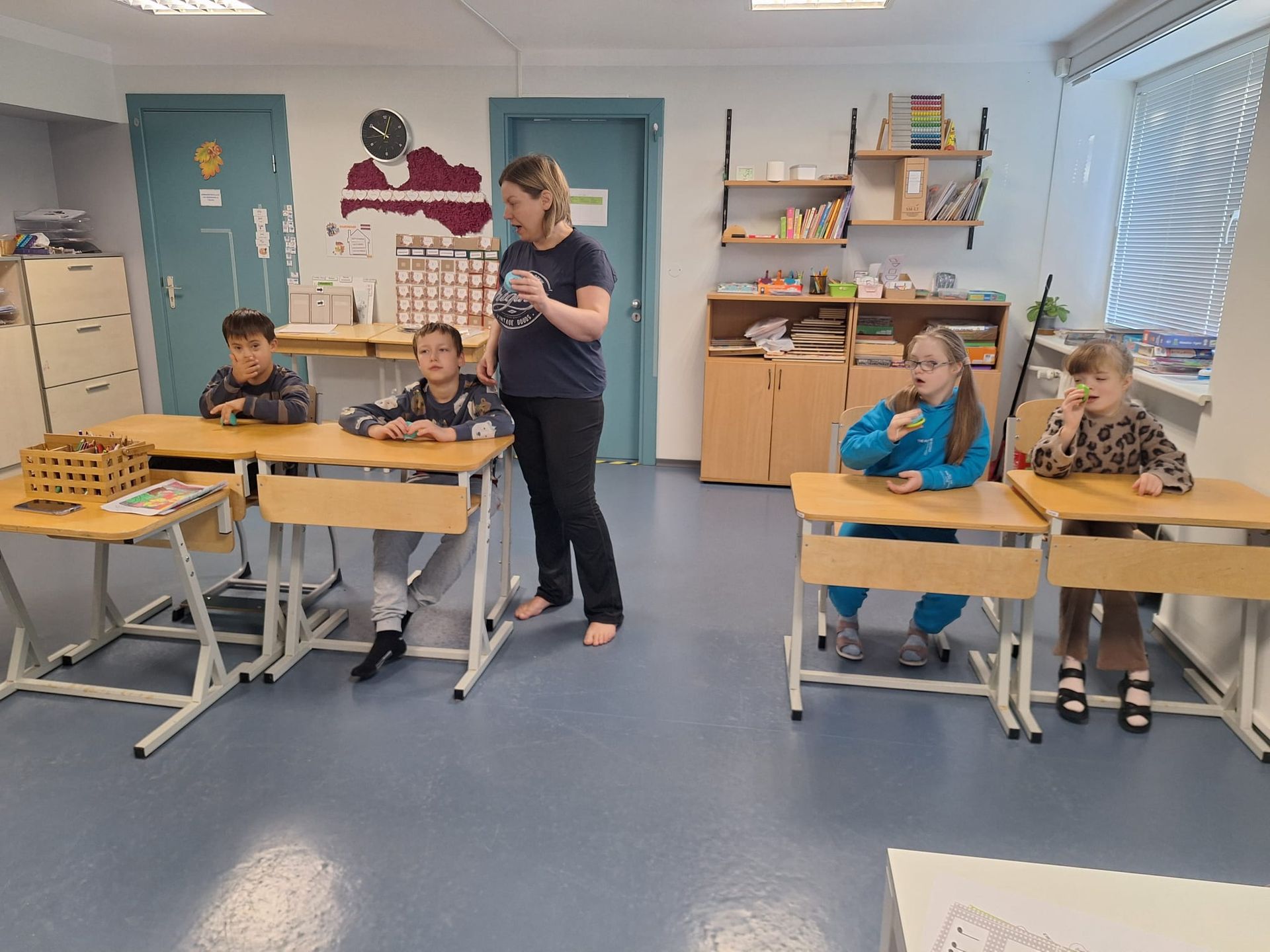 A teacher and four students in a classroom. Students sit at desks. The teacher stands and gestures.