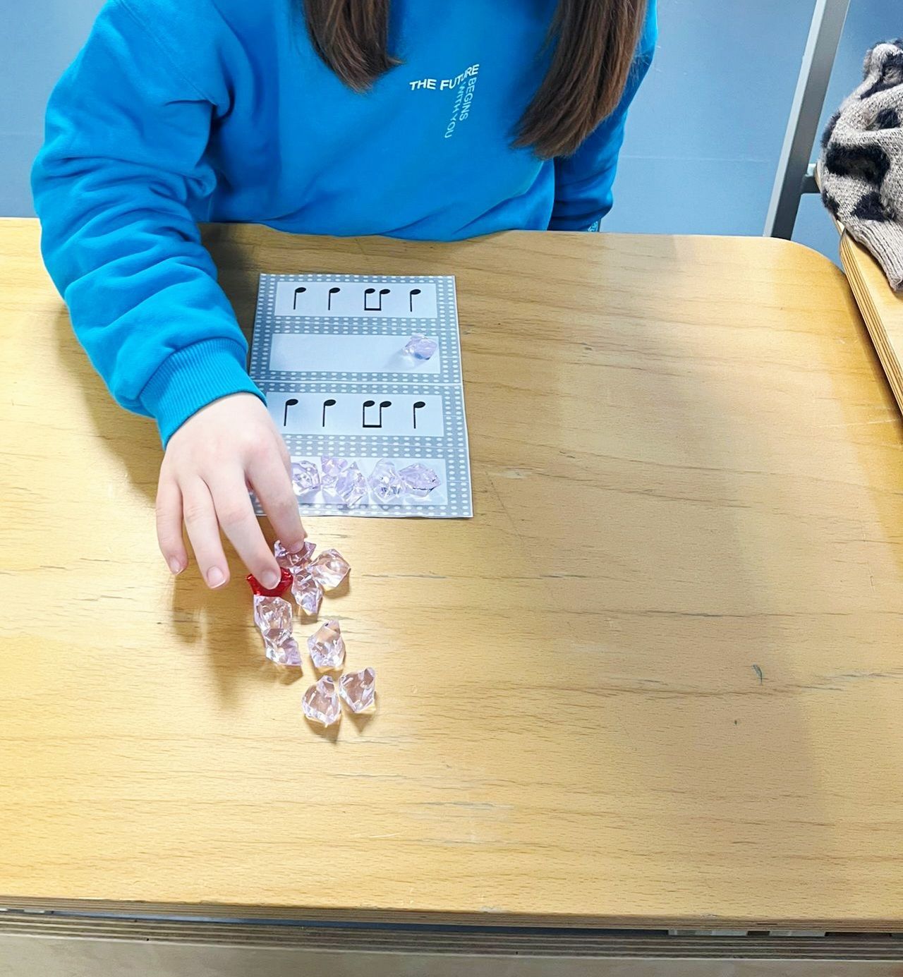 Person's hand placing pink crystals on a music-themed game card on a wooden table. Blue shirt.