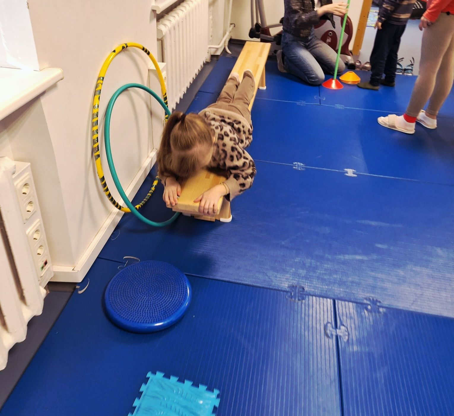 Child lying on a balance beam in a gymnasium, head down, with hula hoops and mats.