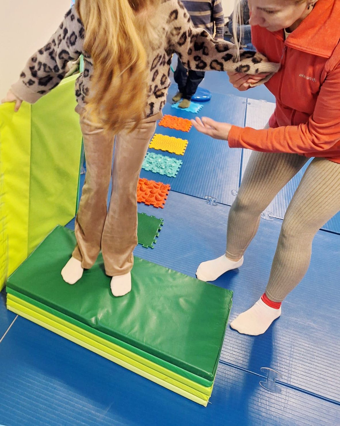 Girl walks on colorful stepping stones, guided by an adult in a gym.