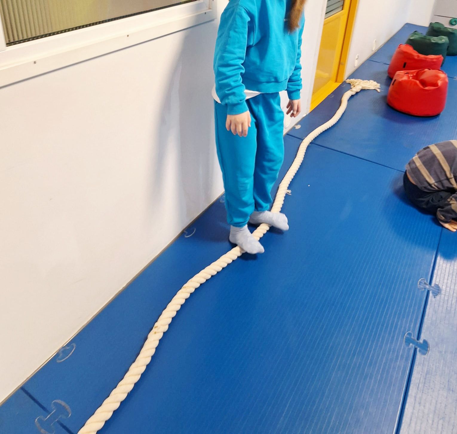 A child in blue sweats walks on a rope laid on a blue mat in a gym.