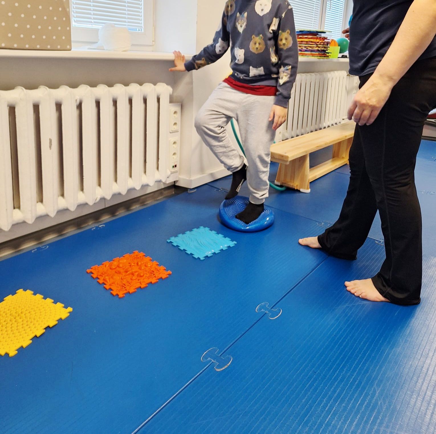 Child balancing on a disc, next to a person, on a blue floor with tactile squares. Therapy setting.