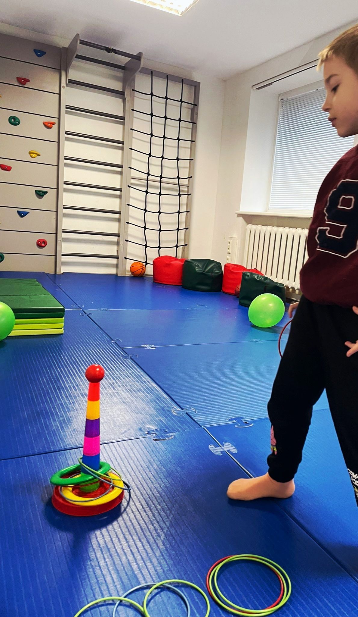 Child playing ring toss on a blue mat. Rings are on the floor. A climbing wall is in the background.