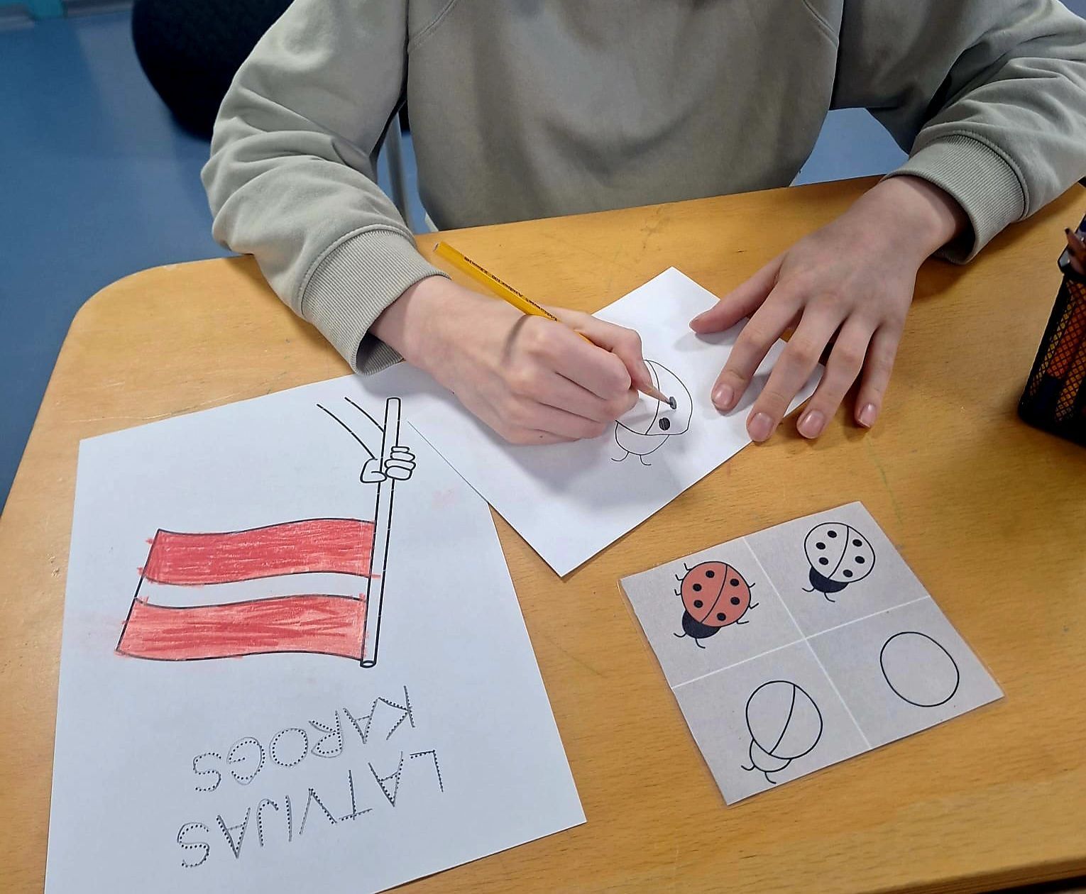 Person drawing on paper with a pencil at a desk with drawings of a flag and ladybugs.