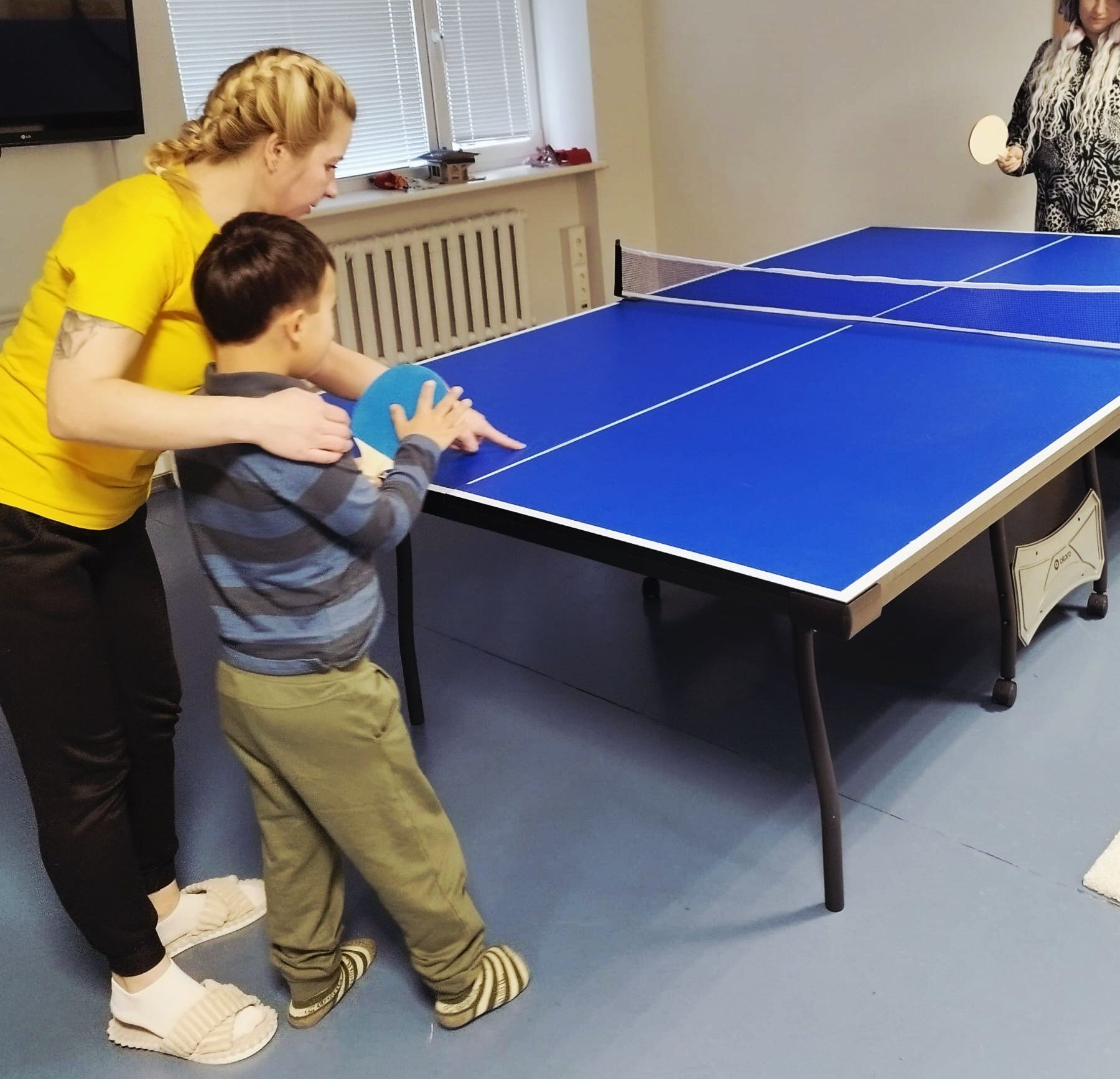 Woman helping a child play table tennis indoors. They are near a blue table, the child holds a paddle.