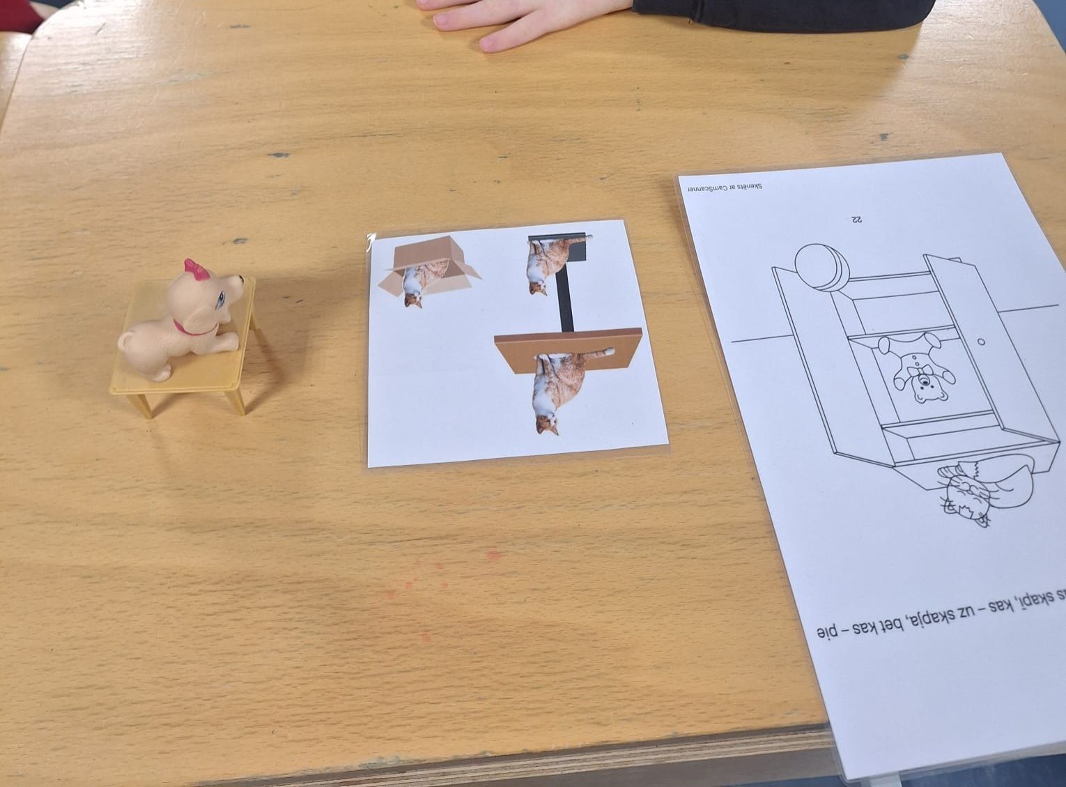 A small toy cat on a chair next to a square paper with upside-down images and a line drawing on white paper.