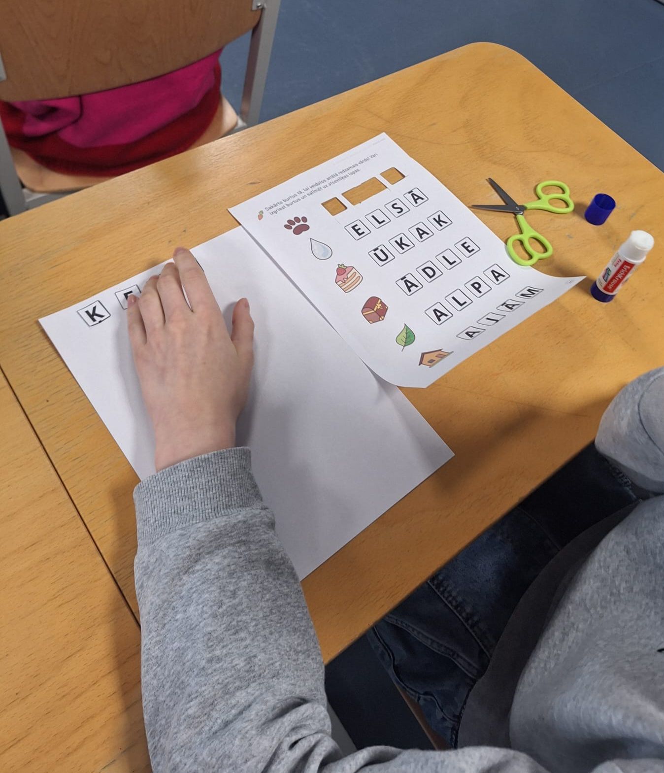 Person's hand on paper, worksheet with words, scissors, glue stick on desk in classroom.