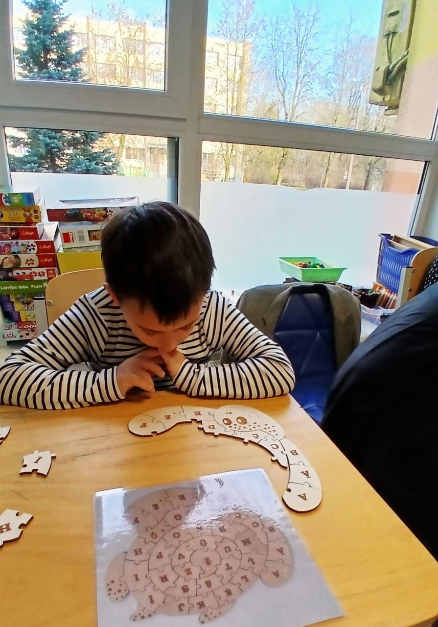 A child sits at a table, looking down at a wooden puzzle. Inside a room, by a window.