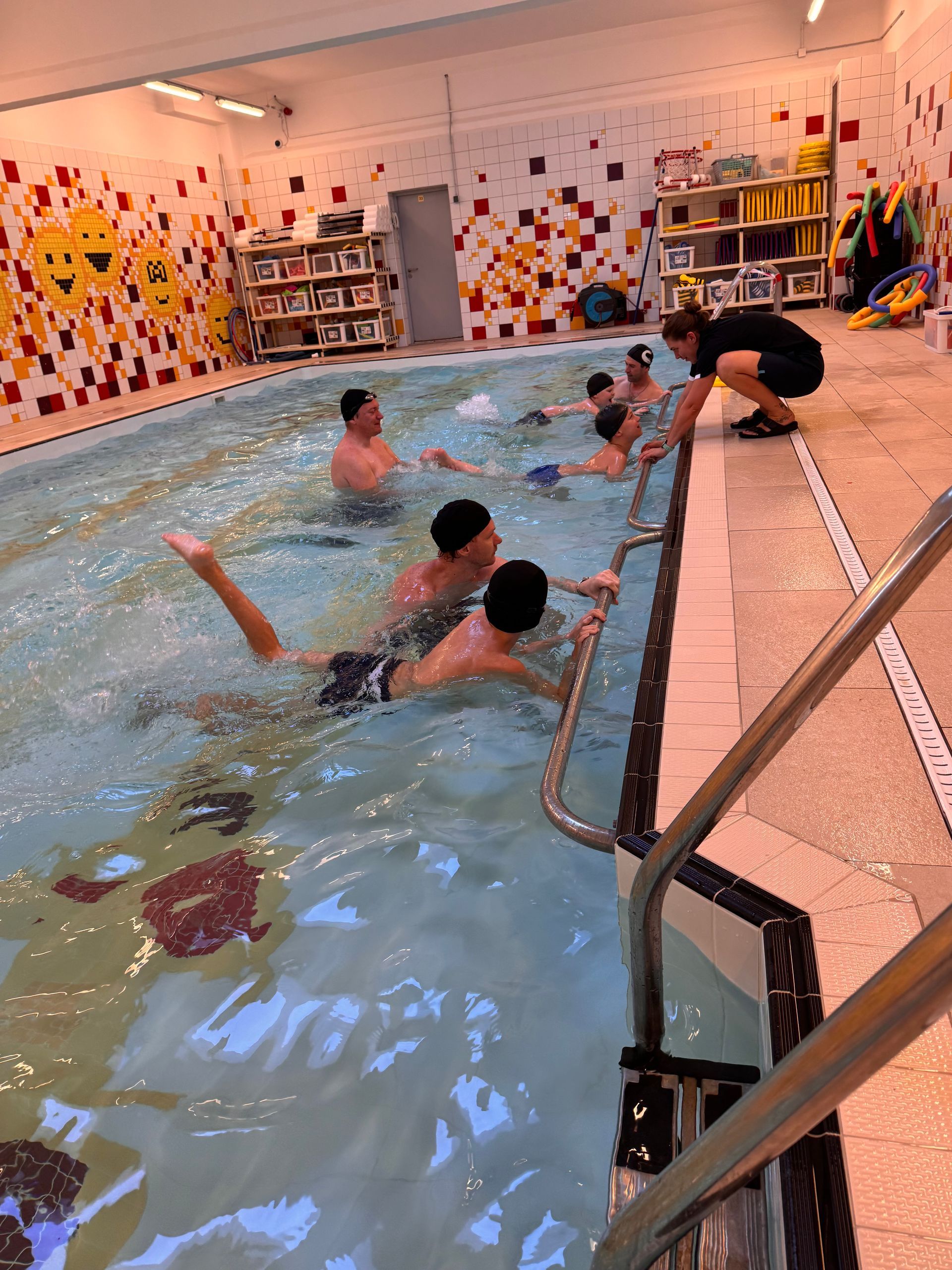 A swimming instructor coaches a group of children in an indoor pool with colorful tiled walls.