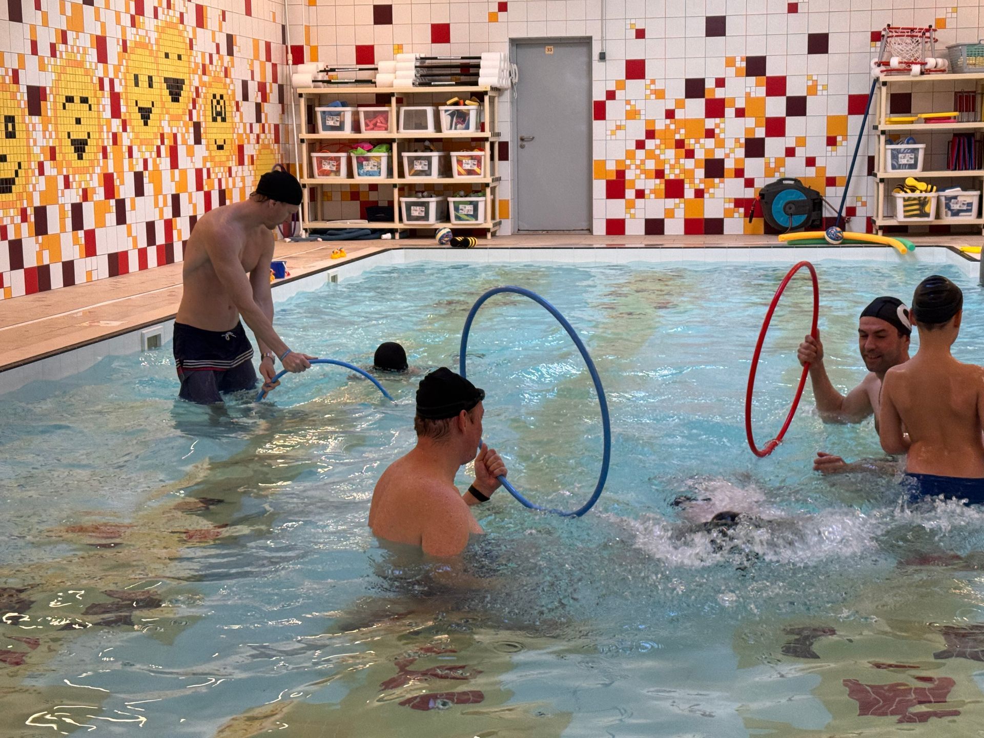 People in a pool interact with hoops, surrounded by a mosaic wall with yellow smiley faces and shelves of supplies.