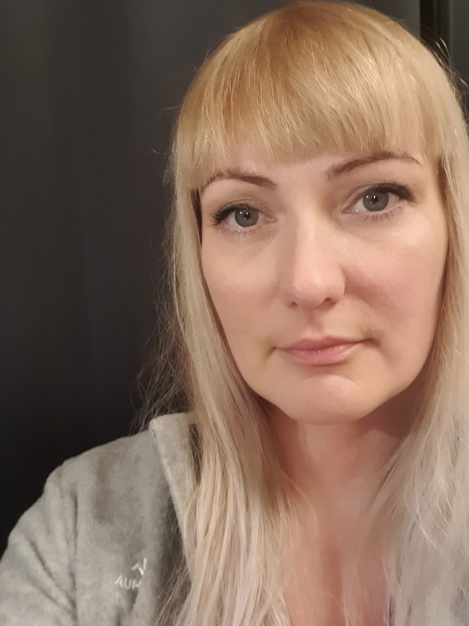 A person with blonde hair and bangs looks toward the camera against a plain dark background, wearing a grey zip-up hoodie.