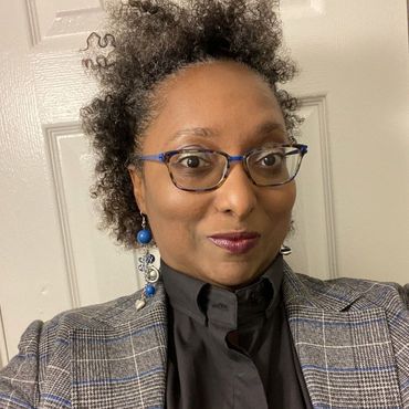 A woman wearing glasses and a jacket is standing in front of a door.
