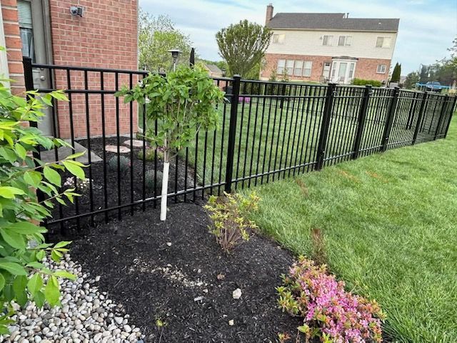 A black fence surrounds a lush green yard with a brick house in the background.