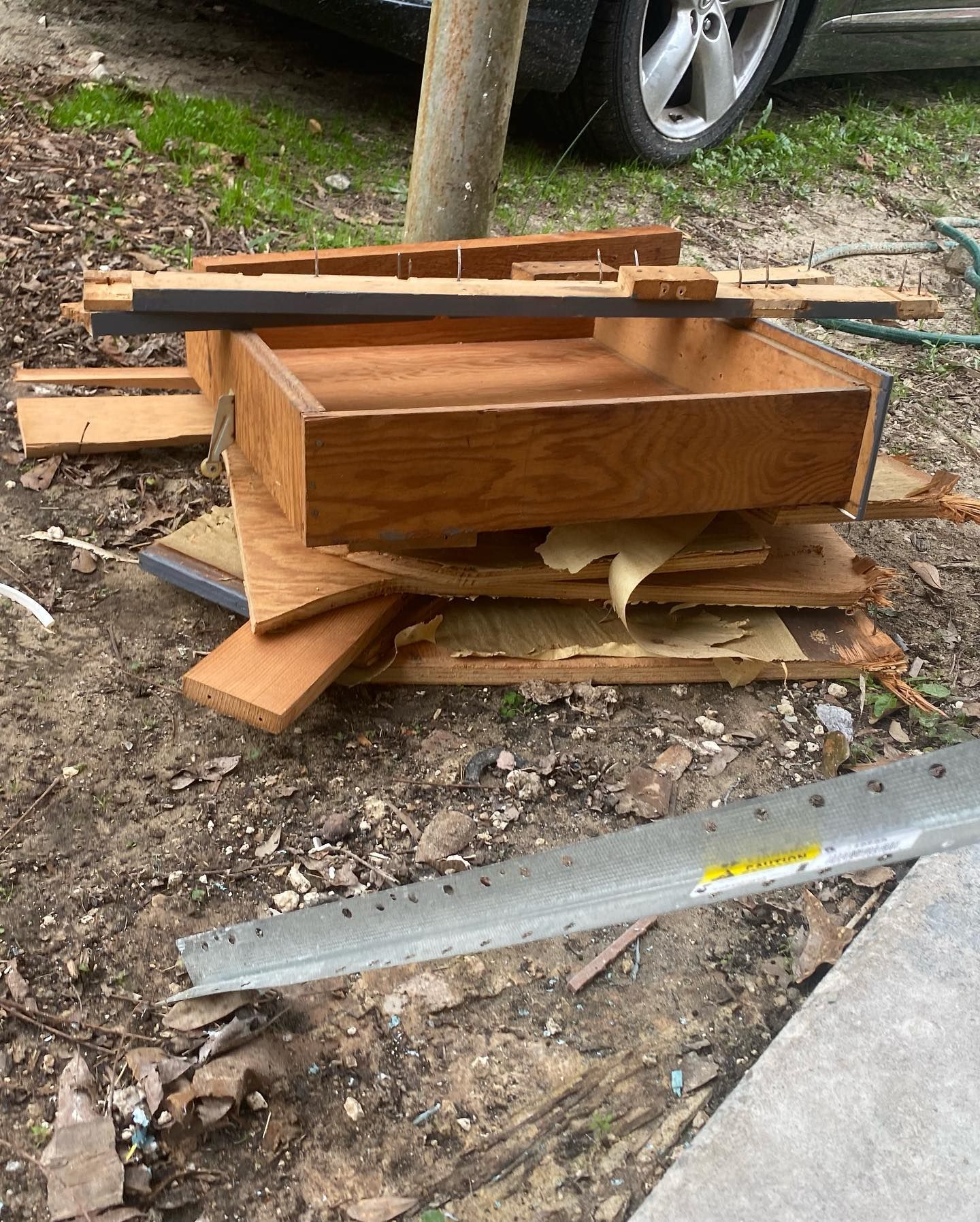 Pile of wood debris, including a drawer, on the ground near a car and a concrete edge.