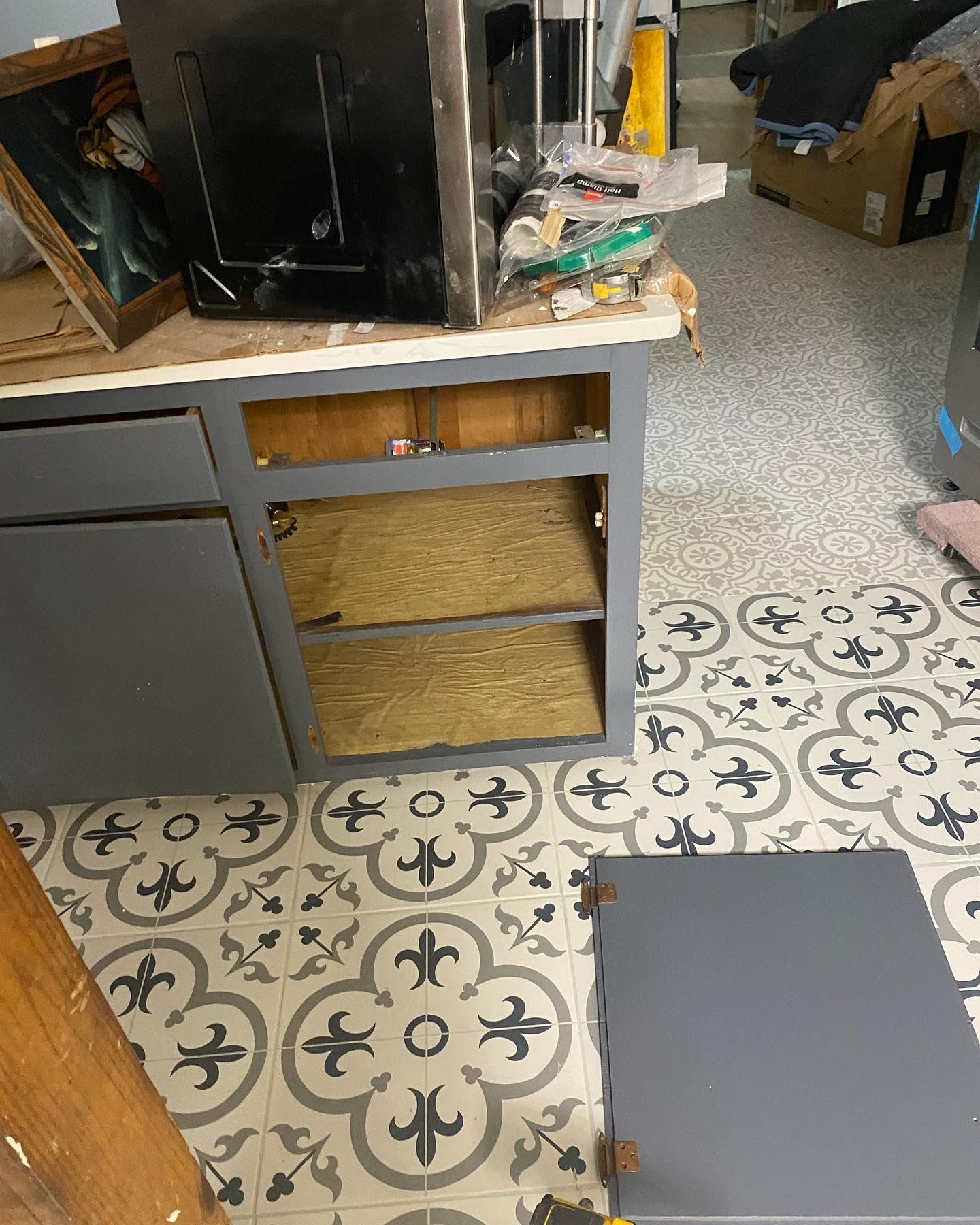 Gray kitchen cabinet with open drawers and a detached door on patterned floor.