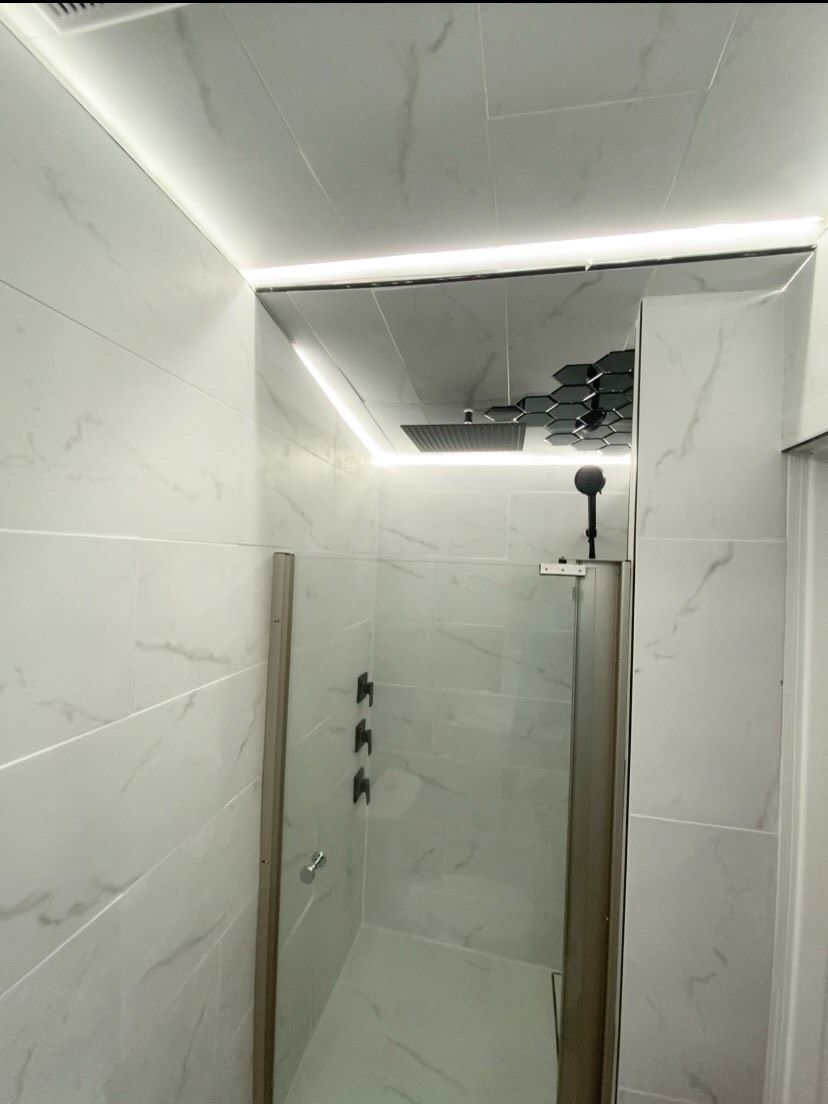 A modern white bathroom with a glass shower enclosure and linear LED lighting.