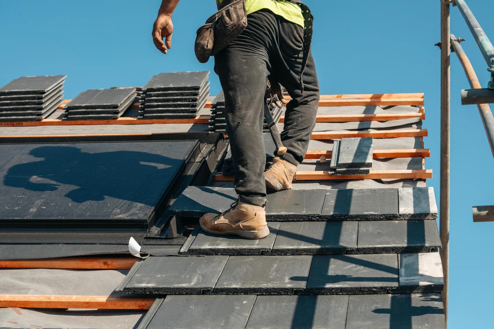 Roofer on a rooftop, installing dark gray tiles against a blue sky.