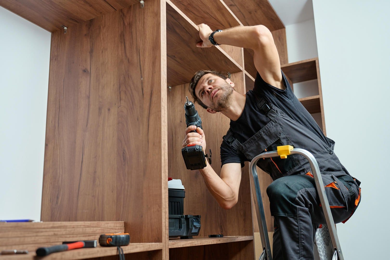 Man on a ladder installing shelves in a wooden closet using a drill.