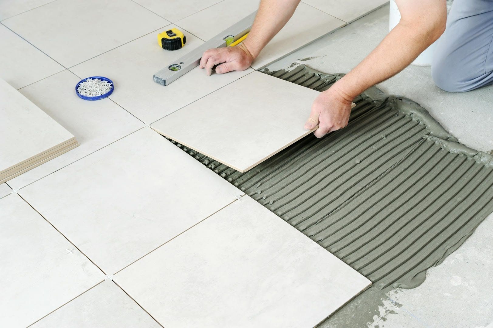 Person installing floor tiles, using a level, trowel, and spacers. Gray cement and beige tiles.