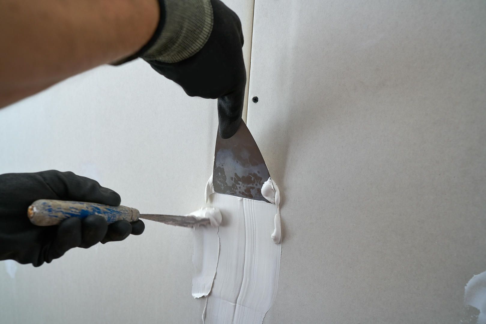 Person wearing gloves using putty knives to apply joint compound to a wall seam.
