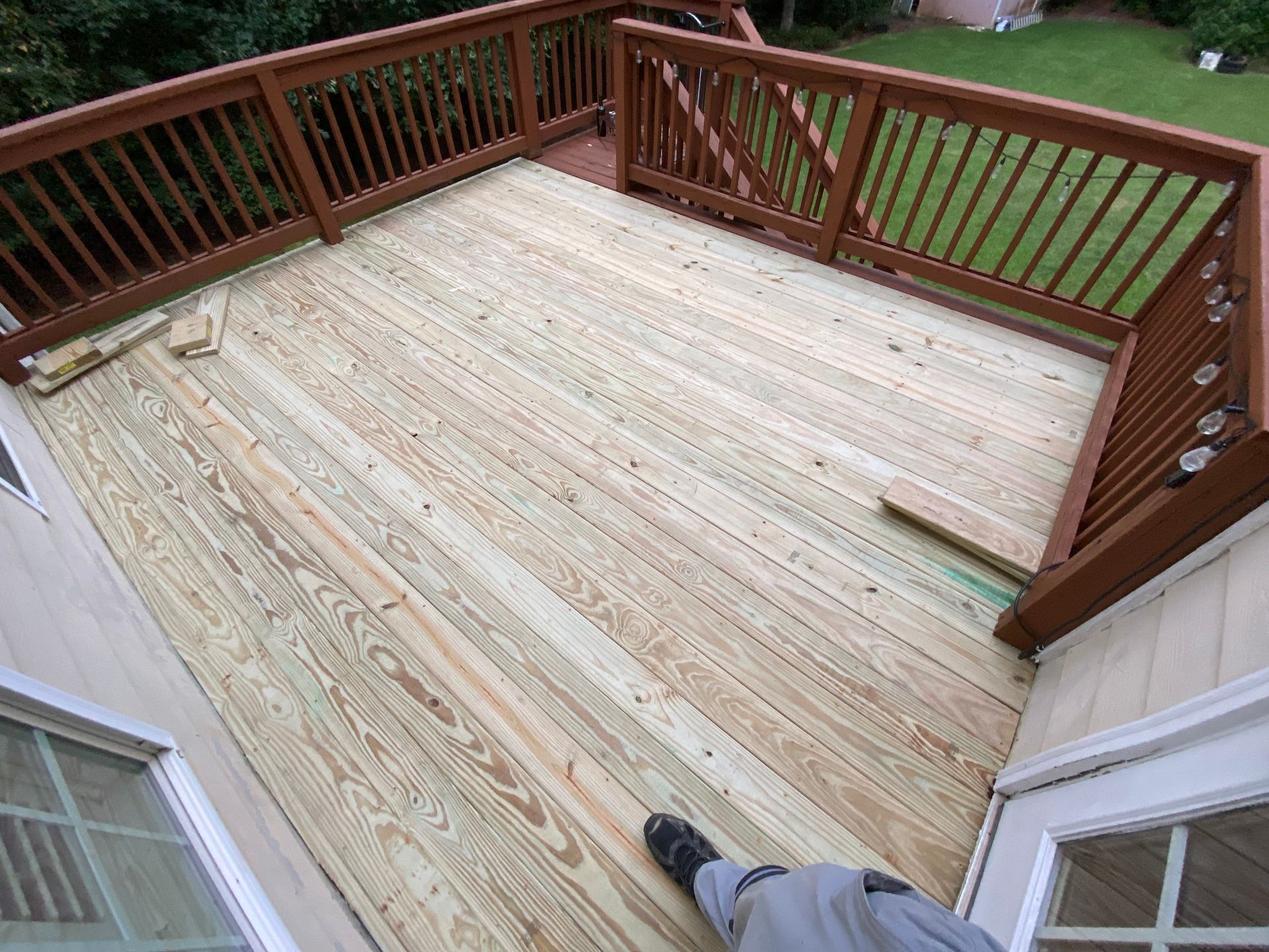 Wooden deck with brown railings, overlooking a grassy yard. A person's legs and foot are visible.
