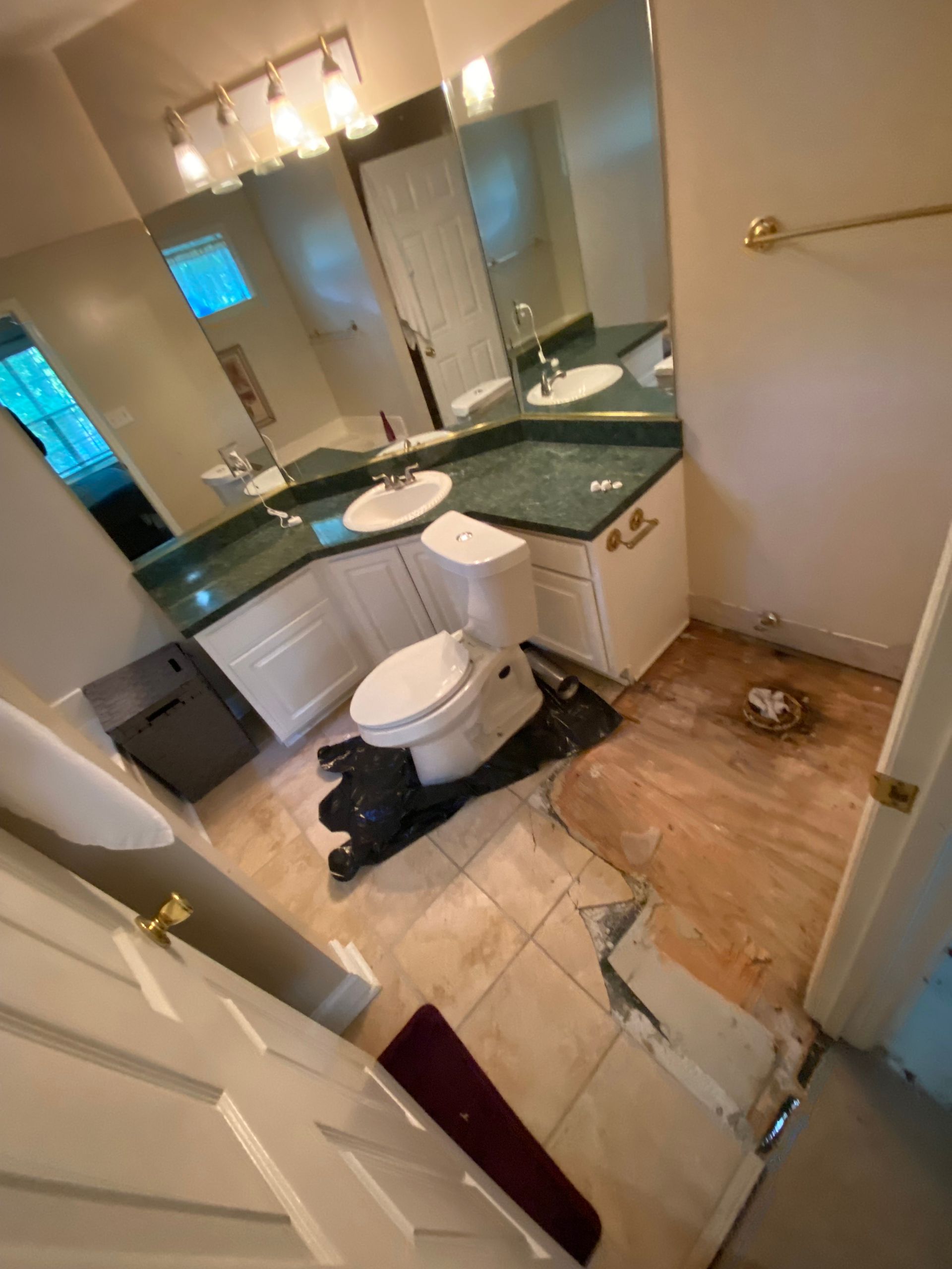 Bathroom renovation in progress, damaged floor, toilet on a black tarp, white cabinets, and green countertops.