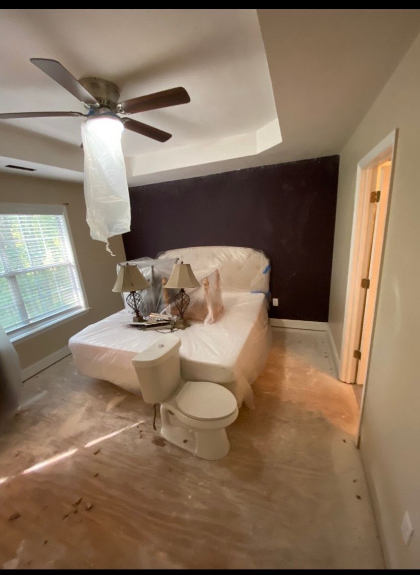 Bedroom with bed and lamps covered in plastic, toilet in front of bed, ceiling fan, dark wall.