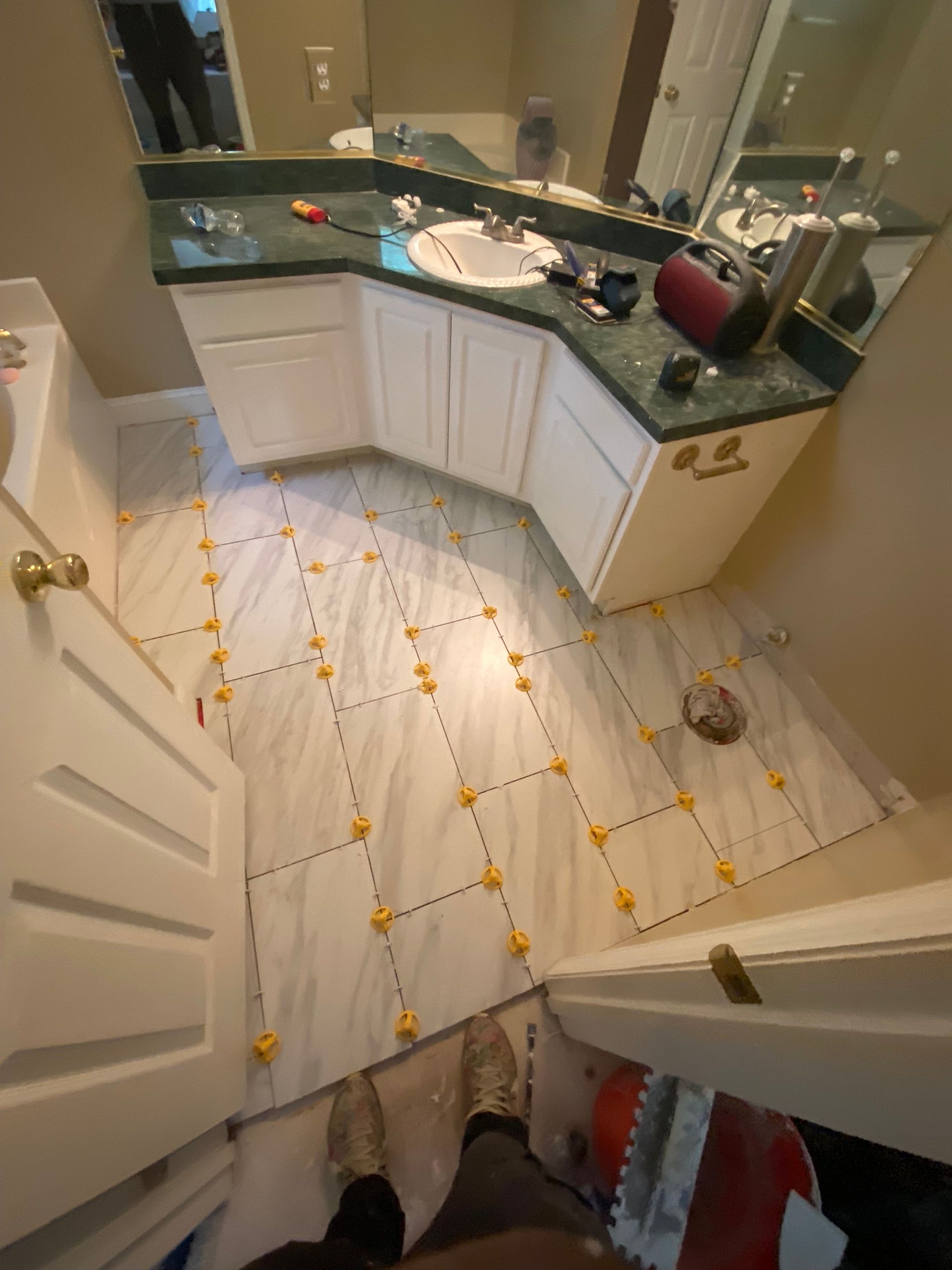 Bathroom floor tiling in progress with white tiles and yellow spacers. Green countertop and white cabinets.
