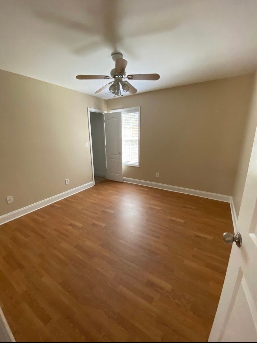 Empty bedroom with wood-look floor, tan walls, ceiling fan, and window with natural light.