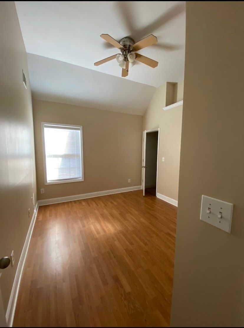 Empty bedroom with vaulted ceiling, hardwood floors, window, and ceiling fan. Beige walls and natural light.