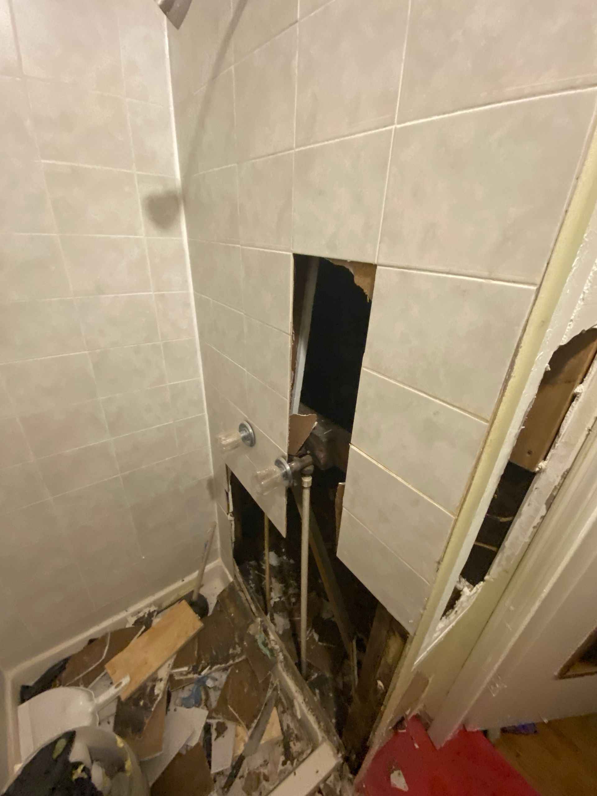 Damaged bathroom wall with exposed pipes and debris. White tiles and drywall are broken.