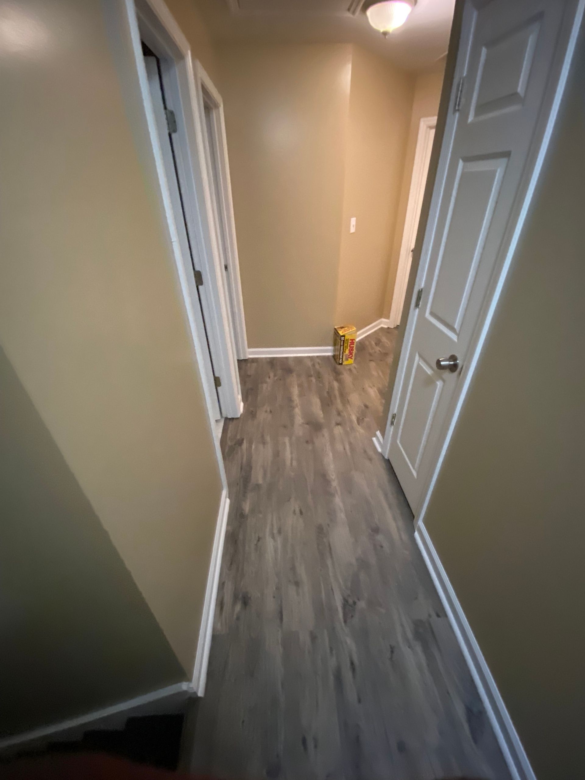 Narrow hallway with beige walls, white trim, and gray wood-look flooring. Three white doors line the hallway.