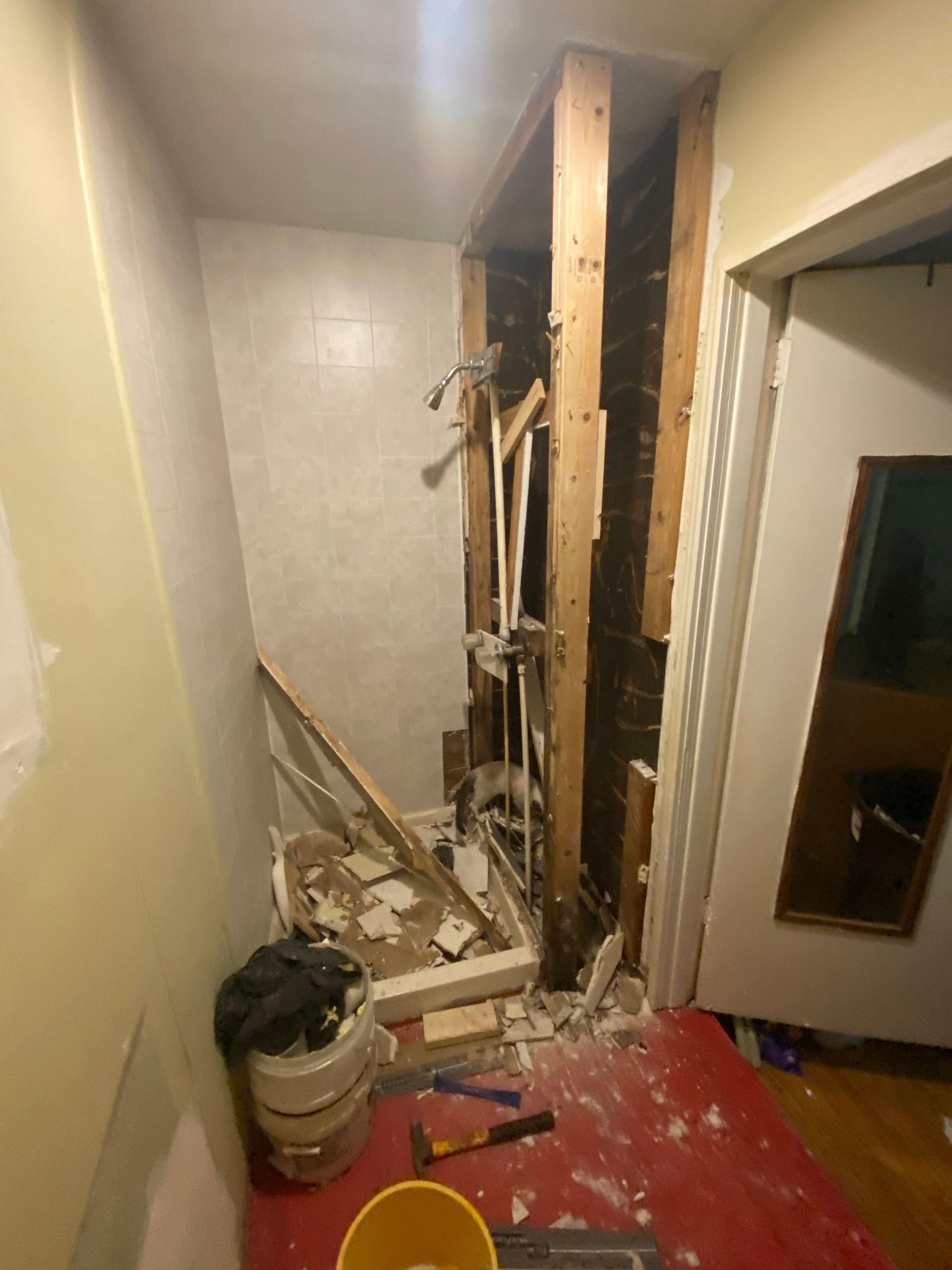 Bathroom renovation in progress, walls torn out exposing wooden studs and plumbing. Debris on floor, tools visible.