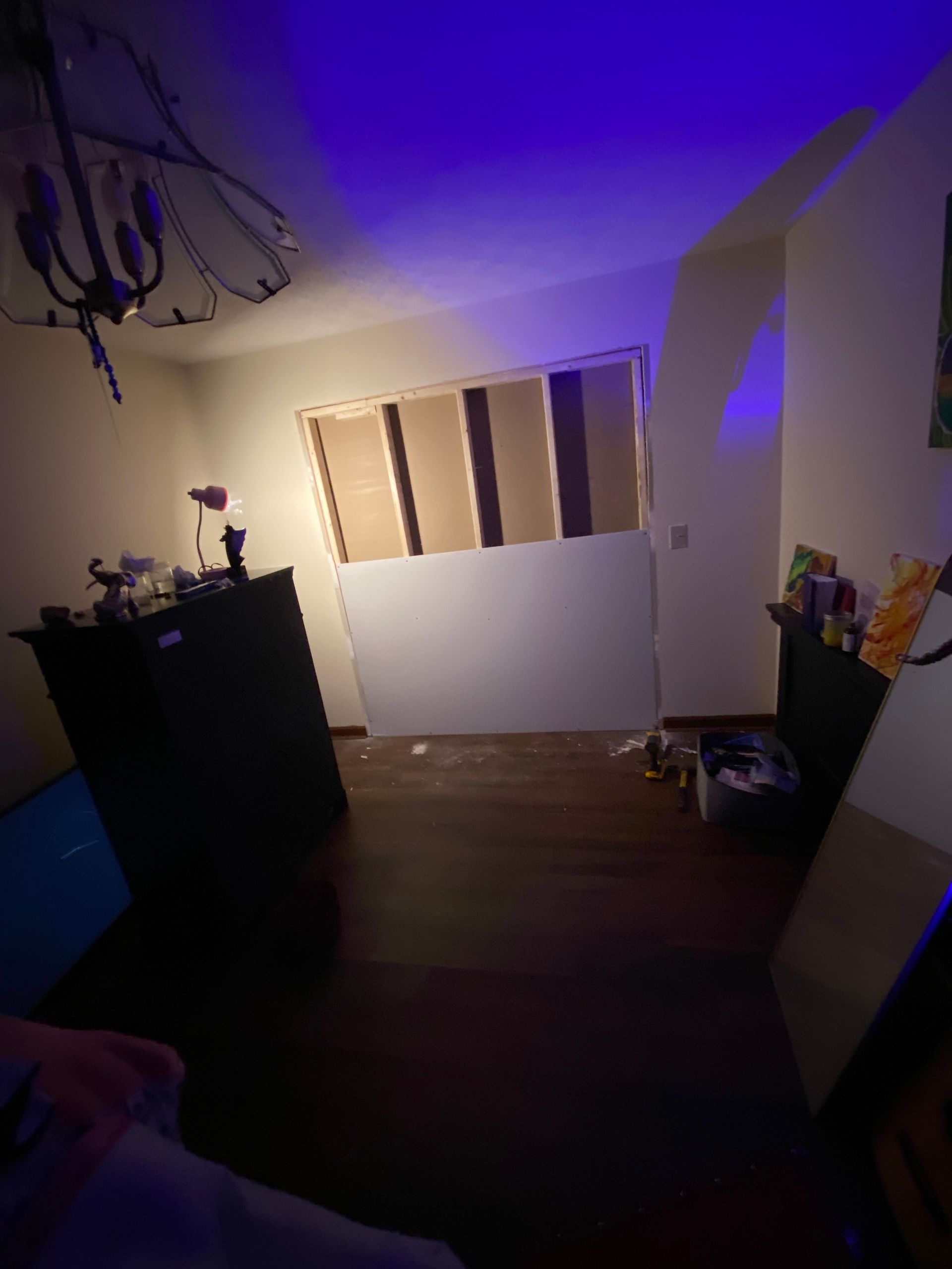 Room with exposed wall studs, drywall, and dark wood floor. Dark dresser and blue lighting.