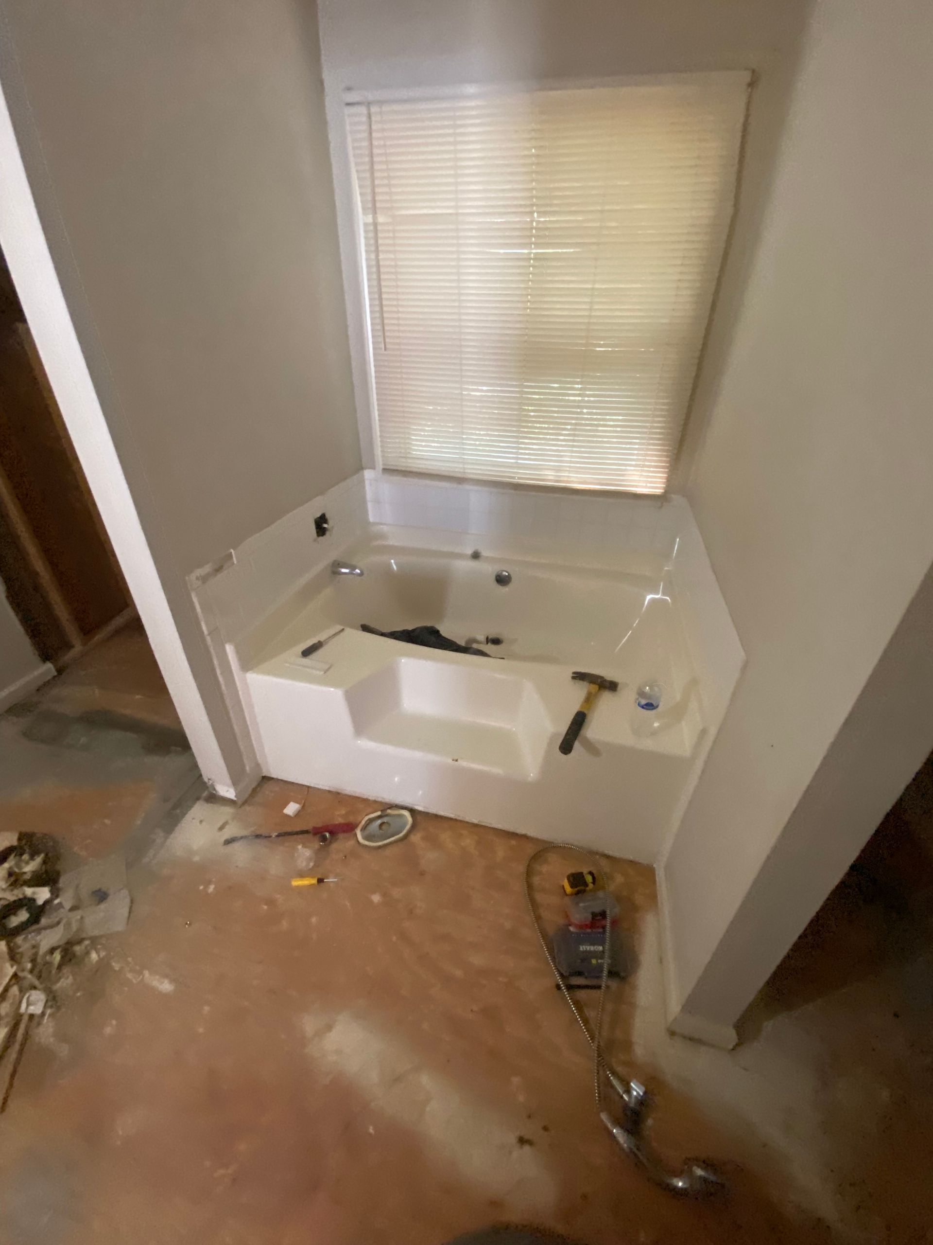 White corner bathtub installed near a window with blinds, construction tools on the floor.