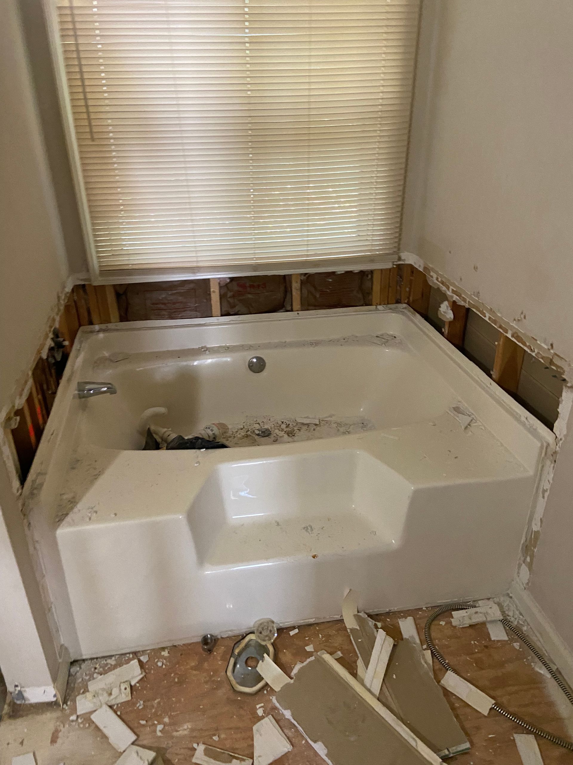 Bathroom with a built-in tub, demolition in progress. Exposed wall studs, debris on floor, blinds in window.