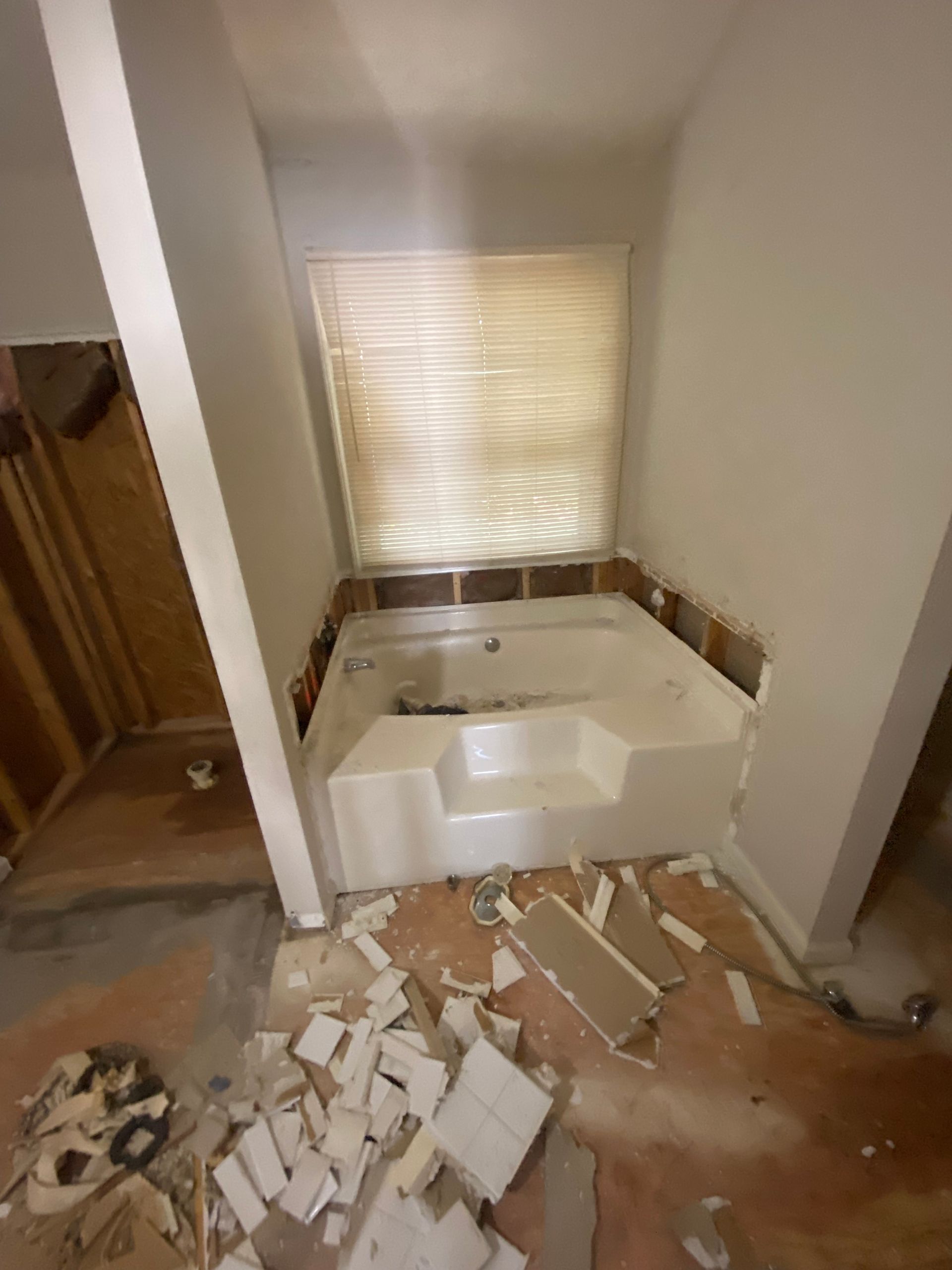 Bathroom renovation, showing a white built-in tub with step, damaged flooring, exposed walls, and a window with blinds.