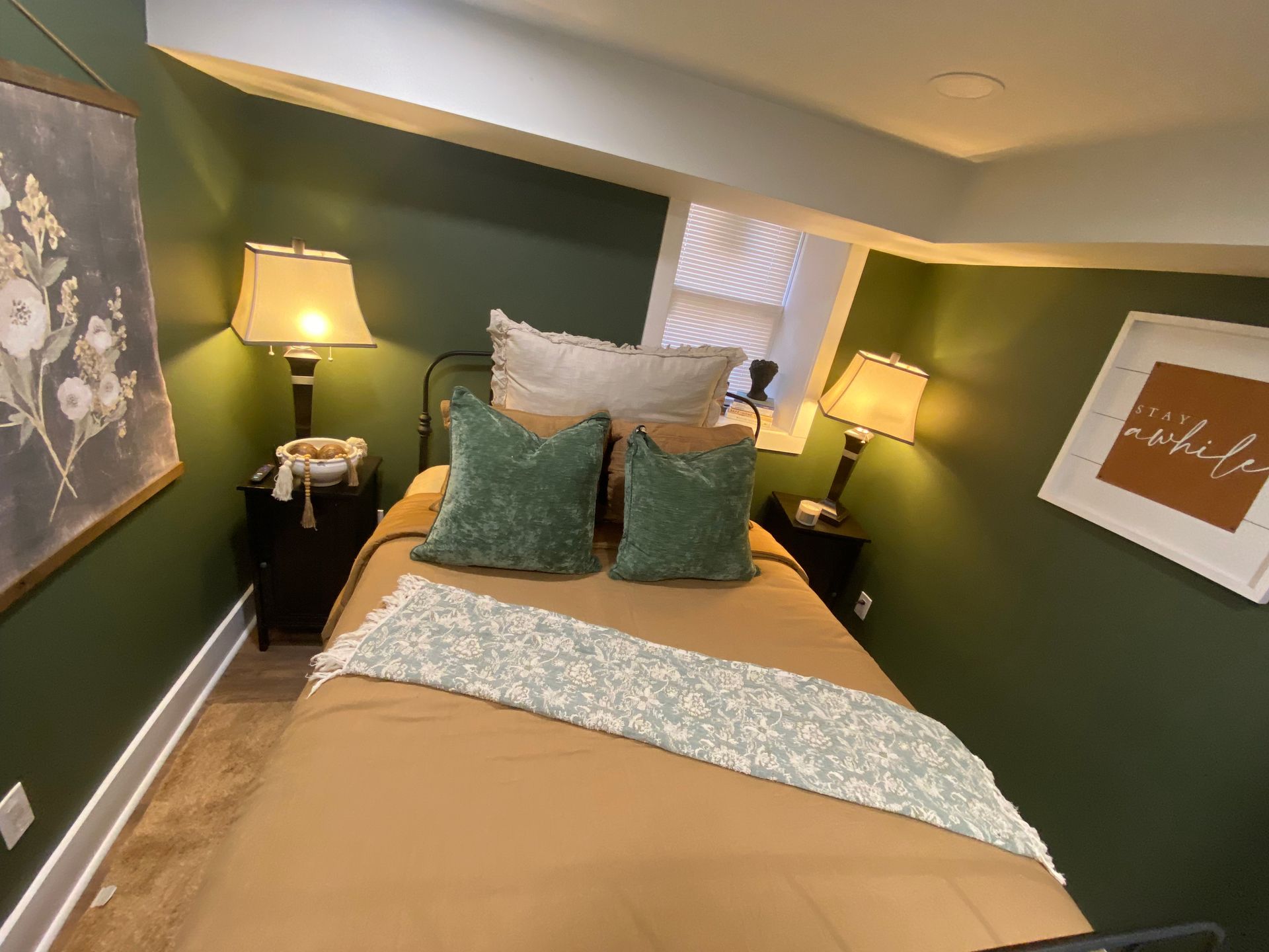 Cozy bedroom with a bed, two lamps, and art on the walls. The walls are green, and the bed has brown sheets.
